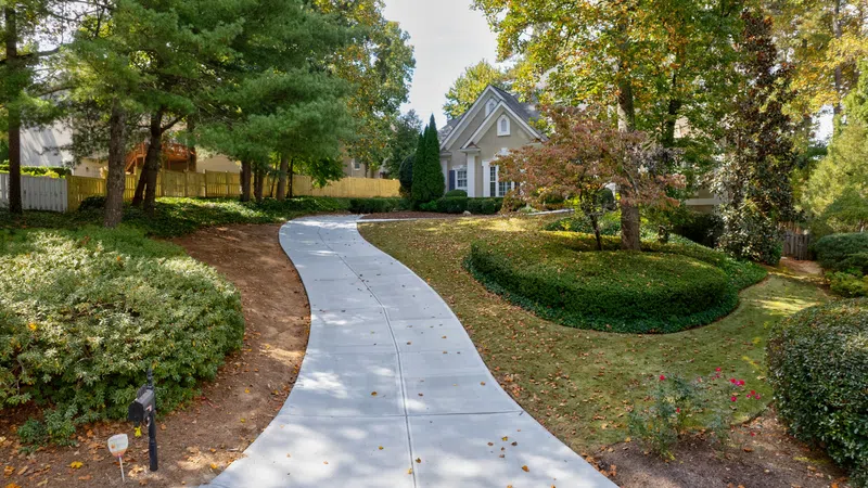 Curved concrete driveway leading to a charming house surrounded by trees and well-maintained landscaping.