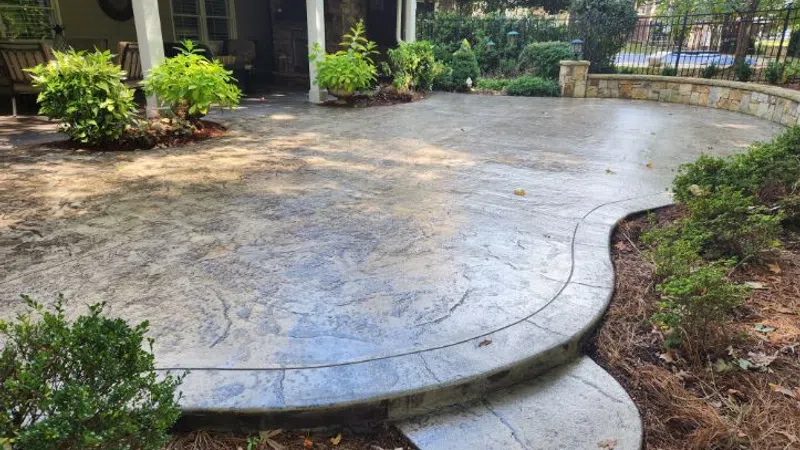 Curved stamped concrete patio with garden beds and plants under a covered porch in a backyard.