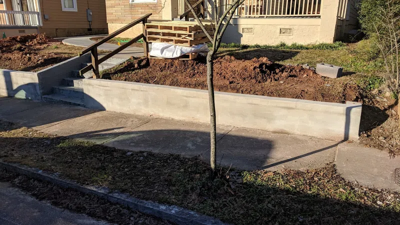 Newly constructed concrete retaining wall and stairs outside a residential house with bare soil and a small tree.