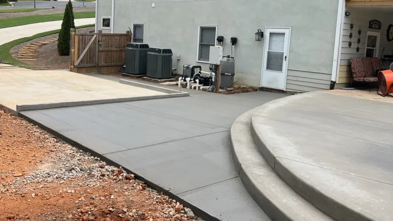 Newly poured concrete driveway and patio beside a modern house, showcasing smooth surfaces and landscaping in view.