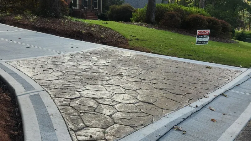 Stamped concrete driveway with a textured pattern in front of a brick house and green lawn.
