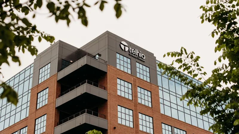 Modern multi-story Telhio Credit Union office building with large windows and balcony, framed by green tree leaves.