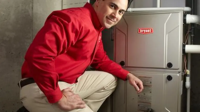 Technician in red shirt kneeling next to a Bryant HVAC system inside a basement with concrete walls.