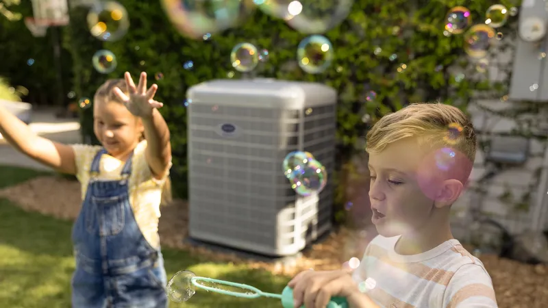 Two children playing with bubbles in a sunny backyard near an outdoor air conditioning unit.
