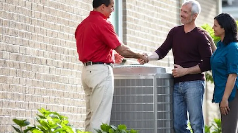Technician shaking hands with couple by a newly installed home air conditioning unit outside their brick house.