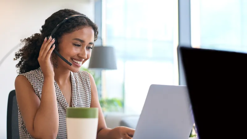 Smiling woman with headset working on laptop at a bright office desk with coffee cup nearby