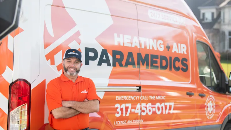 Man in orange uniform stands confidently next to a service van for Heating + Air Paramedics.