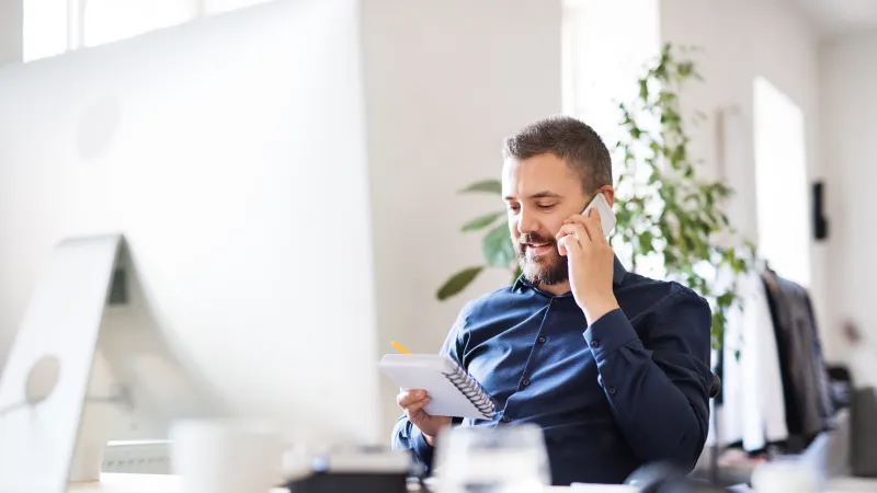 Man in blue shirt talking on phone and taking notes in a bright modern office with plants and computer.