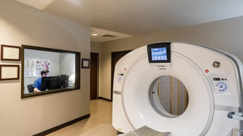 Modern CT scan machine in a clinical setting with staff at a computer in the background.