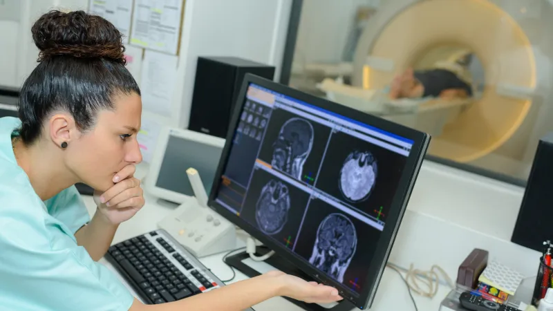 A medical professional examines MRI scans on a computer while a patient undergoes a scan in the background.