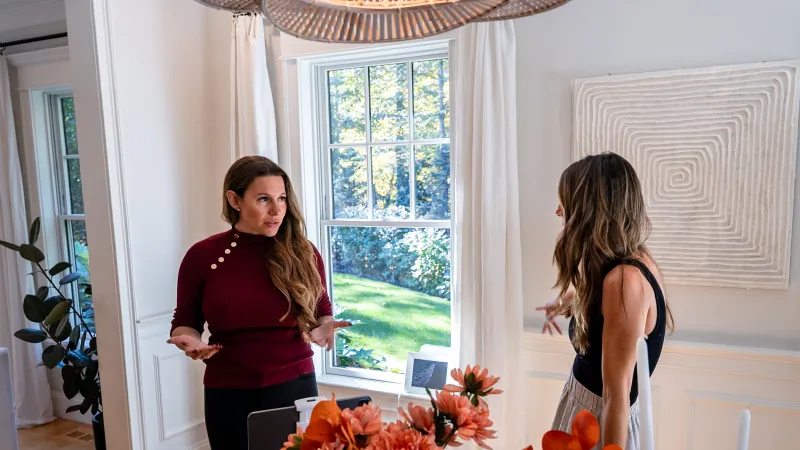 Two women having a discussion in a bright room with a window and a vase of orange flowers on the table.
