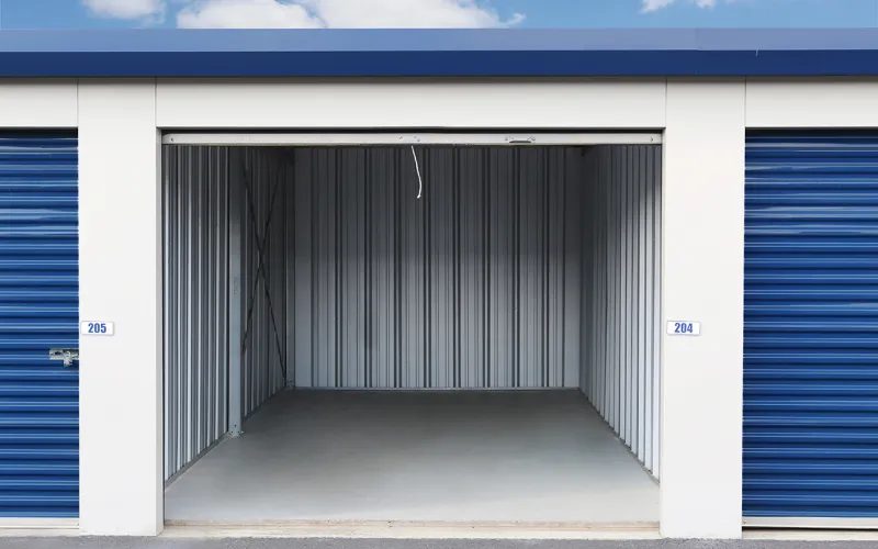 Empty storage unit with gray metal walls and concrete floor between two closed blue roll-up doors under blue sky.