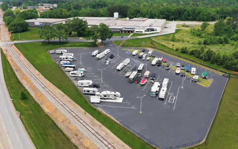 Aerial view of a spacious RV parking lot with various vehicles, surrounded by green trees and an industrial building.