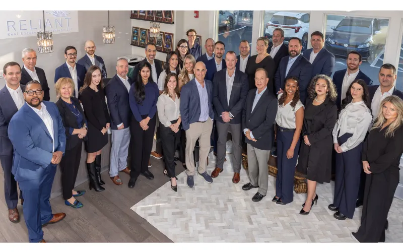Professional diverse real estate team posing in office with awards plaques and Reliant Real Estate logo on wall.