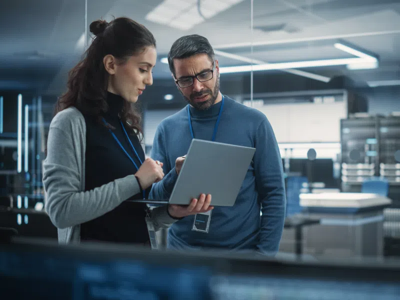 Two IT professionals discuss data on a laptop in a modern server room with networking equipment racks.