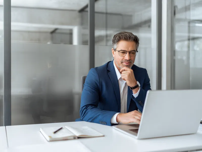 Professional man in blue suit working on laptop at modern office desk with notebook and pen.
