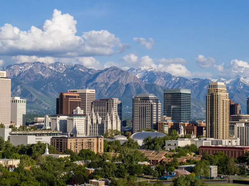 Salt Lake City skyline with tall buildings and snow-capped mountains under a bright blue sky with clouds