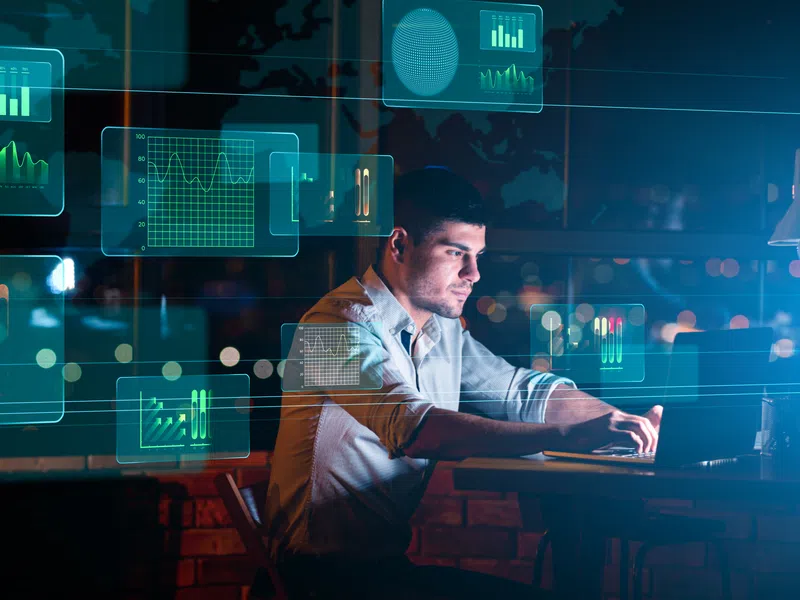 Man working on laptop at night surrounded by digital data and analytics charts floating in the air.