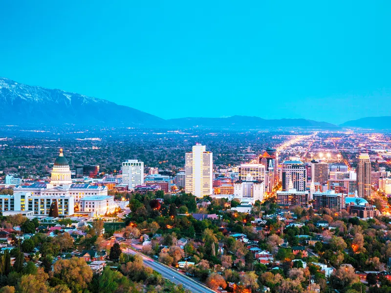 Evening cityscape of Salt Lake City with illuminated buildings and mountains in the background under clear blue sky.