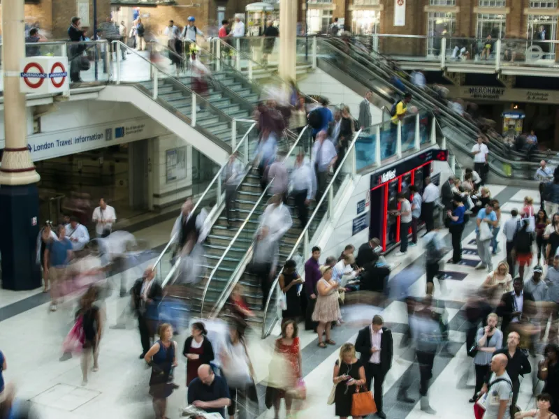 Crowded London train station with people walking, escalators, and travel information signs bustling with activity.