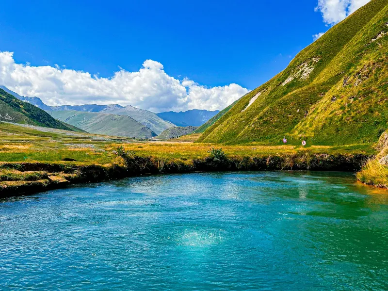 Clear blue pond surrounded by grassy hills under a bright blue sky with clouds in a mountainous landscape.