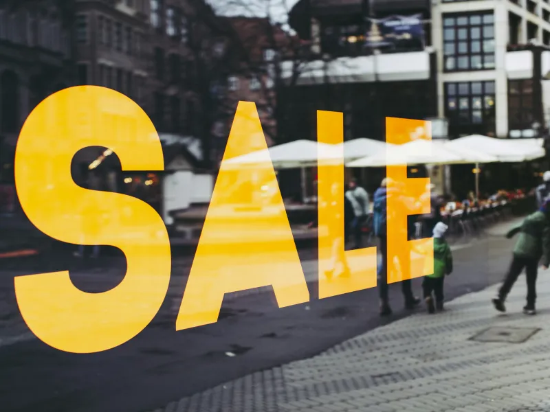 Large yellow SALE sign on shop window reflecting a busy urban street with people and outdoor seating.