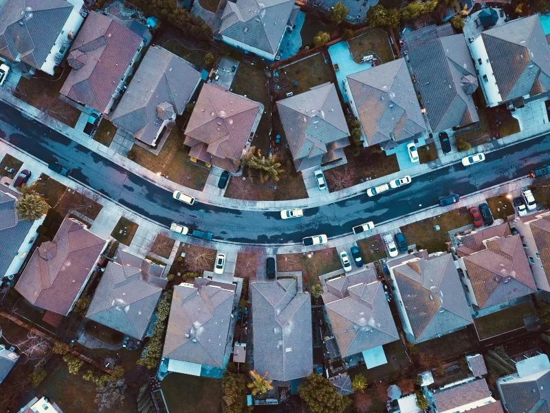 Aerial view of a suburban neighborhood showing houses, streets, yards, and parked cars during dusk.