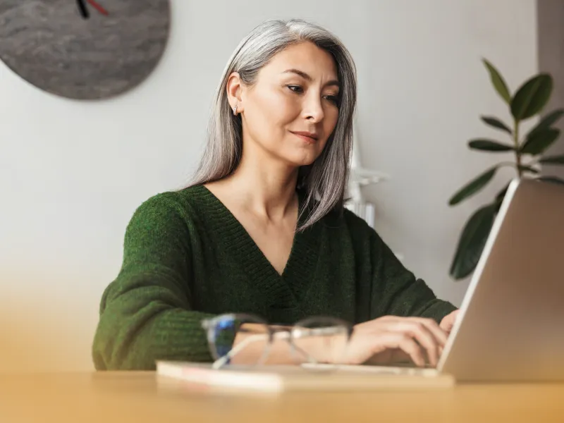 Middle-aged woman with gray hair working on a laptop at a wooden desk with glasses and clock in background