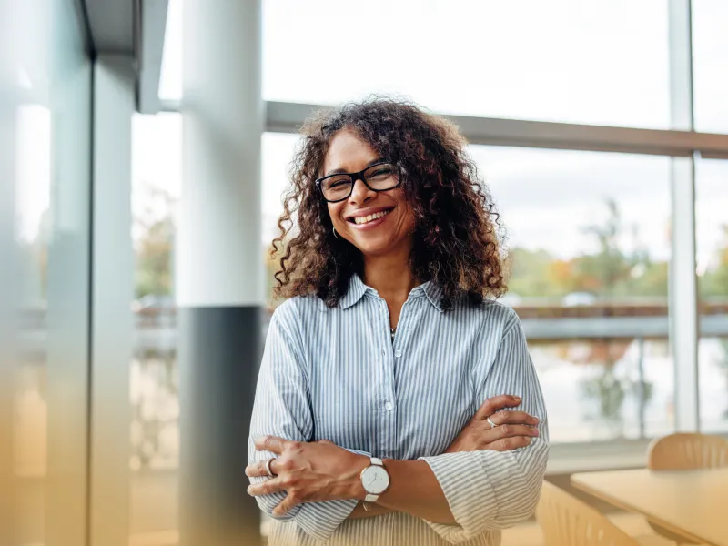 Smiling woman with curly hair and glasses standing in bright modern office with arms crossed.