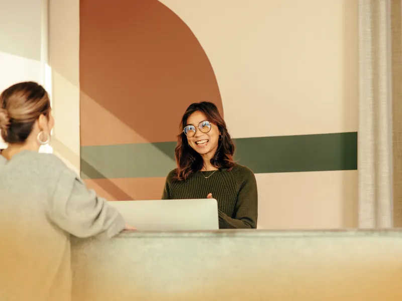 Smiling receptionist in glasses greets a visitor at a modern front desk with colorful geometric wall art.