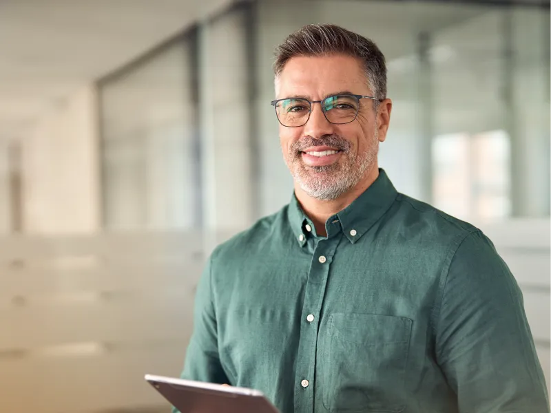 Smiling middle-aged man with glasses holding a tablet in a modern office setting wearing a green button-up shirt