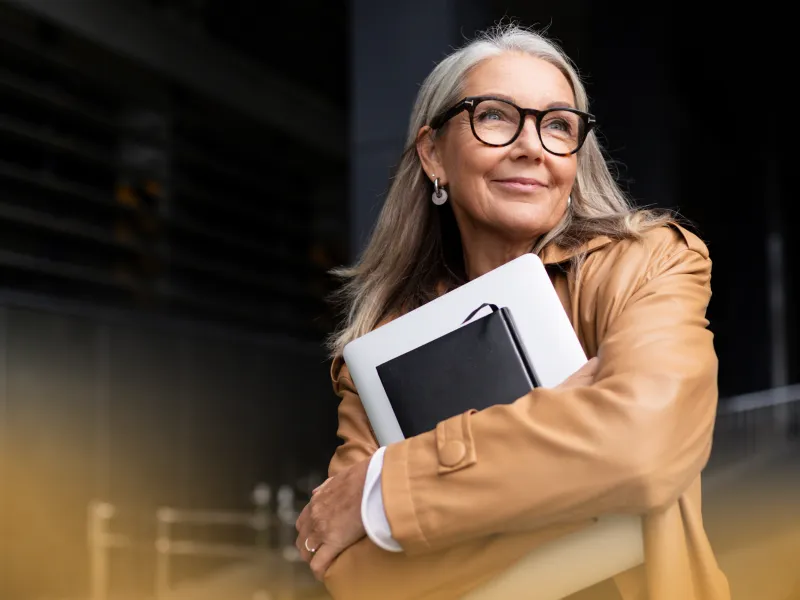 Confident mature woman in glasses and tan coat holding laptop and notebook outdoors, looking thoughtfully sideways.
