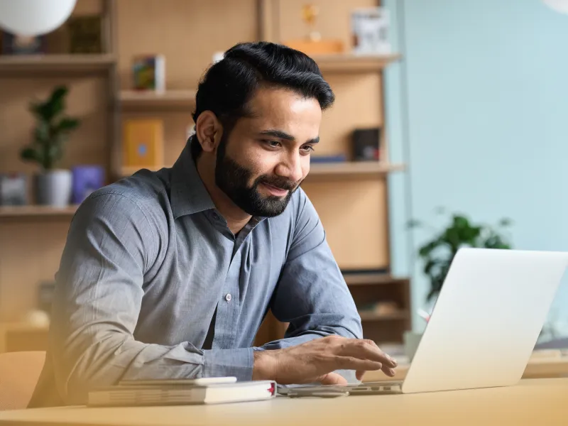 Man in gray shirt working on laptop at a desk in a cozy, modern office with shelves and plants.