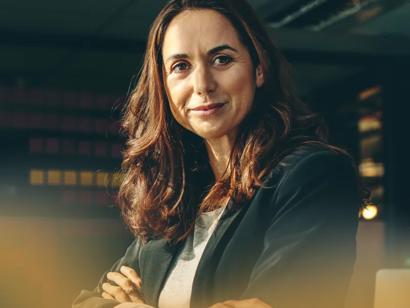 Confident woman with crossed arms wearing a black blazer and smiling in a modern office setting.