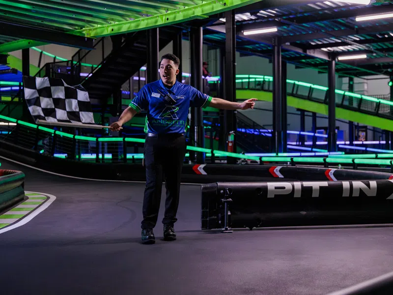 Race official waving a checkered flag at an indoor go-kart track with neon green and blue lighting.