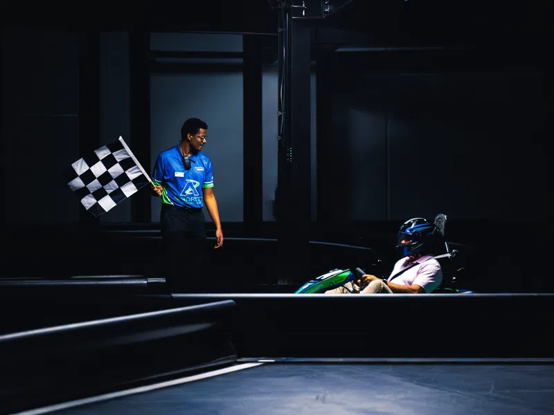 Race official waves checkered flag to finish go-kart race with driver in helmet at indoor track.