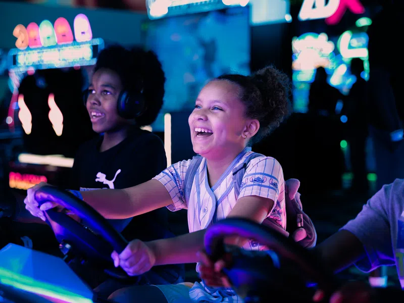 Children smiling and playing an arcade racing game in a vibrant, colorful gaming center with neon lights.