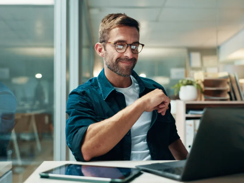 Smiling man wearing glasses working on a laptop in a modern office with tablet on the desk.