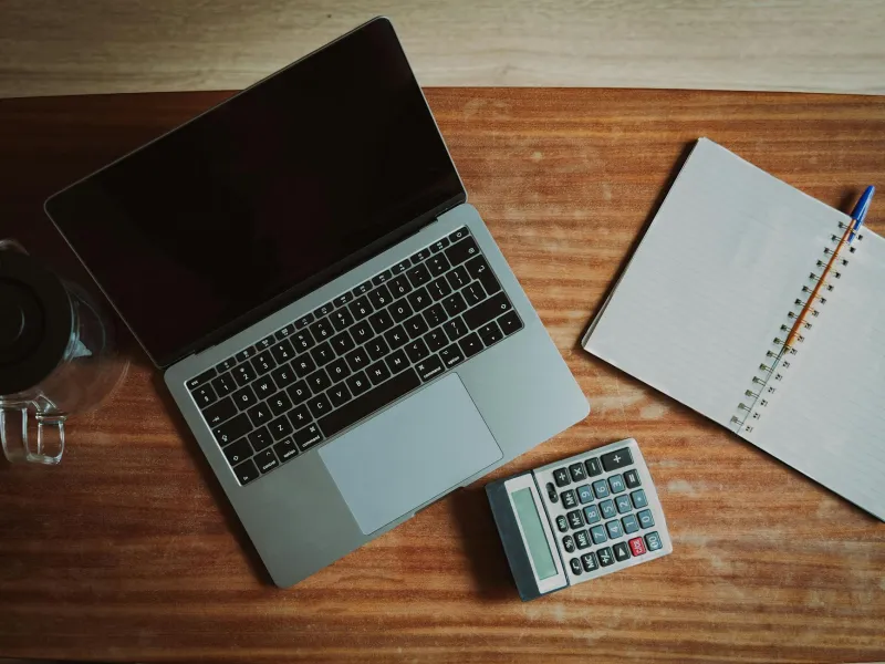 Overhead view of a laptop, calculator, notebook with pen, and glass mug on a wooden desk surface.