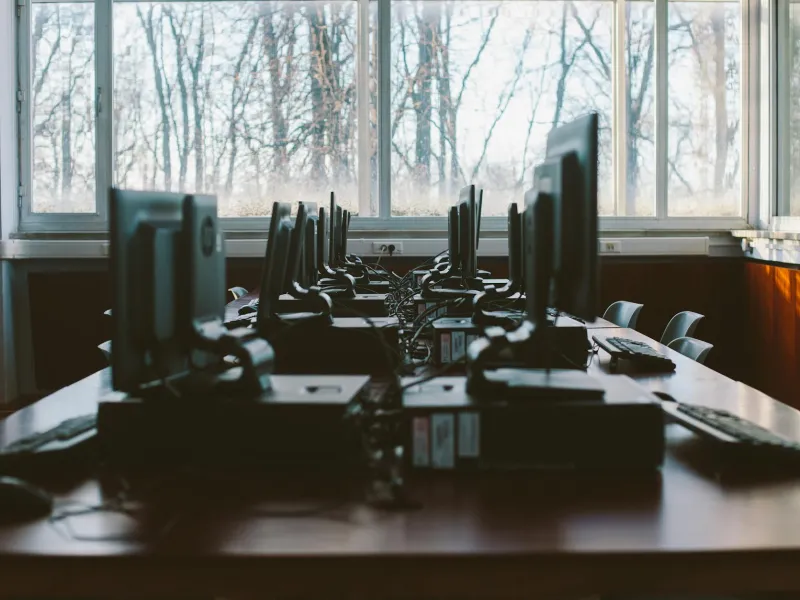 Empty computer lab with rows of desktop PCs on wooden tables and large windows showing leafless trees outside.