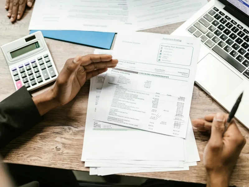Person reviewing bank statements with calculator and laptop on wooden desk for financial planning.