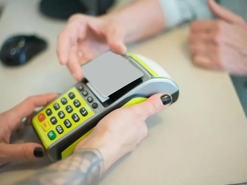 Close-up of a person holding a payment terminal while another person taps a card for contactless payment.