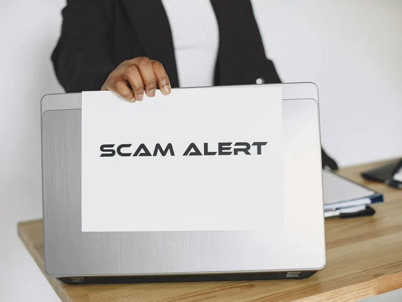 Person holding a sign reading Scam Alert in front of a laptop on a wooden desk.