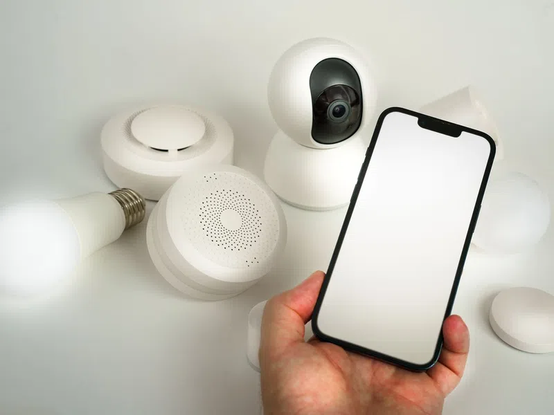 Hand holding smartphone with blank screen next to smart home devices including light bulb, camera, and sensors
