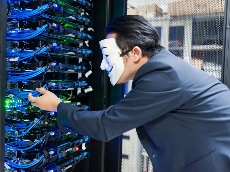 Person wearing Guy Fawkes mask manipulating cables in a server room with network equipment and blue lights