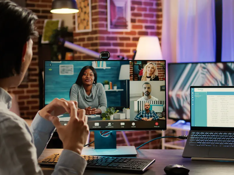 Man attending a virtual video conference call on desktop monitor with four colleagues visible on screen.