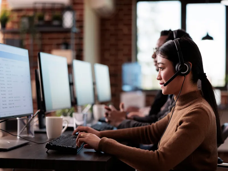 Young woman wearing headset working on computer in modern office with multiple monitors and coworkers.
