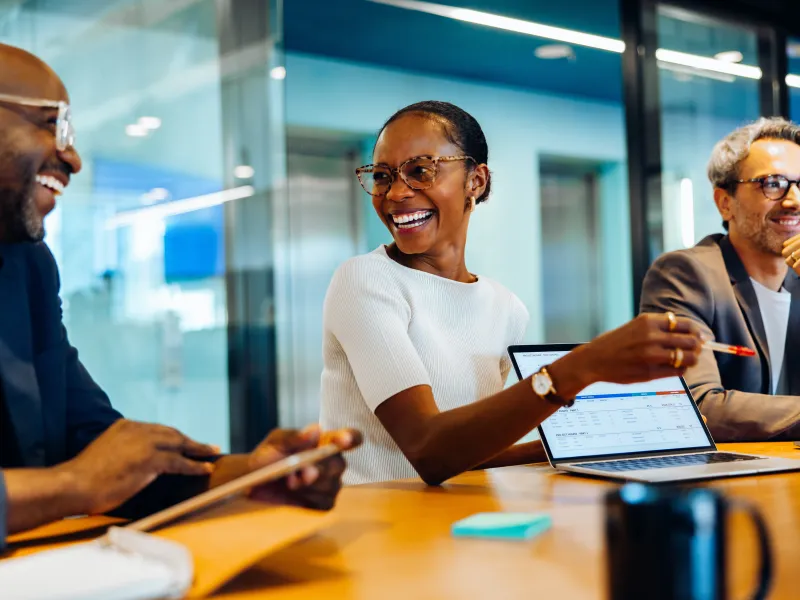 Three professionals smiling and collaborating around a table with digital devices in a modern office.