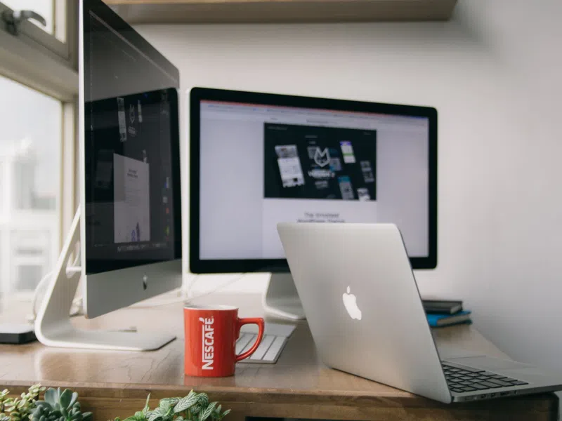 Workstation setup with Apple laptop, iMac, Nescafé red mug, and dual monitors on a wooden desk near window.