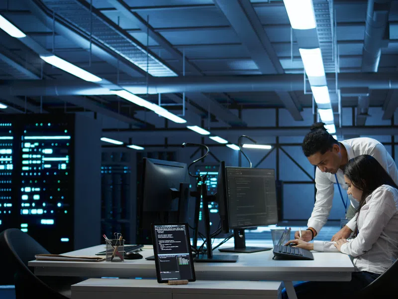 Two IT professionals working late in a server room with multiple monitors and data servers.